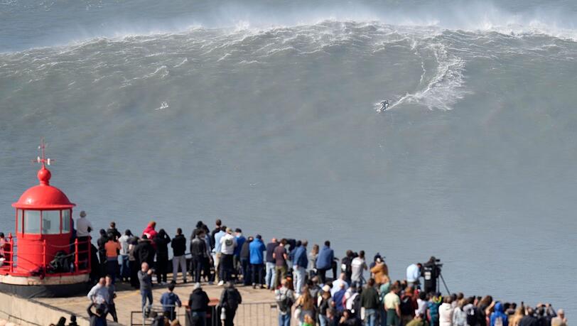 Menschen am Leuchtturm bei Nazaré beobachten einen Surfer am Praia do Norte, der eine Riesenwelle surft. Menschen am Leuchtturm bei Nazaré beobachten einen Surfer am Praia do Norte, der eine Riesenwelle surft.